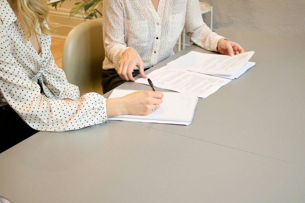 Woman signing on white printer paper beside woman about to touch the documents